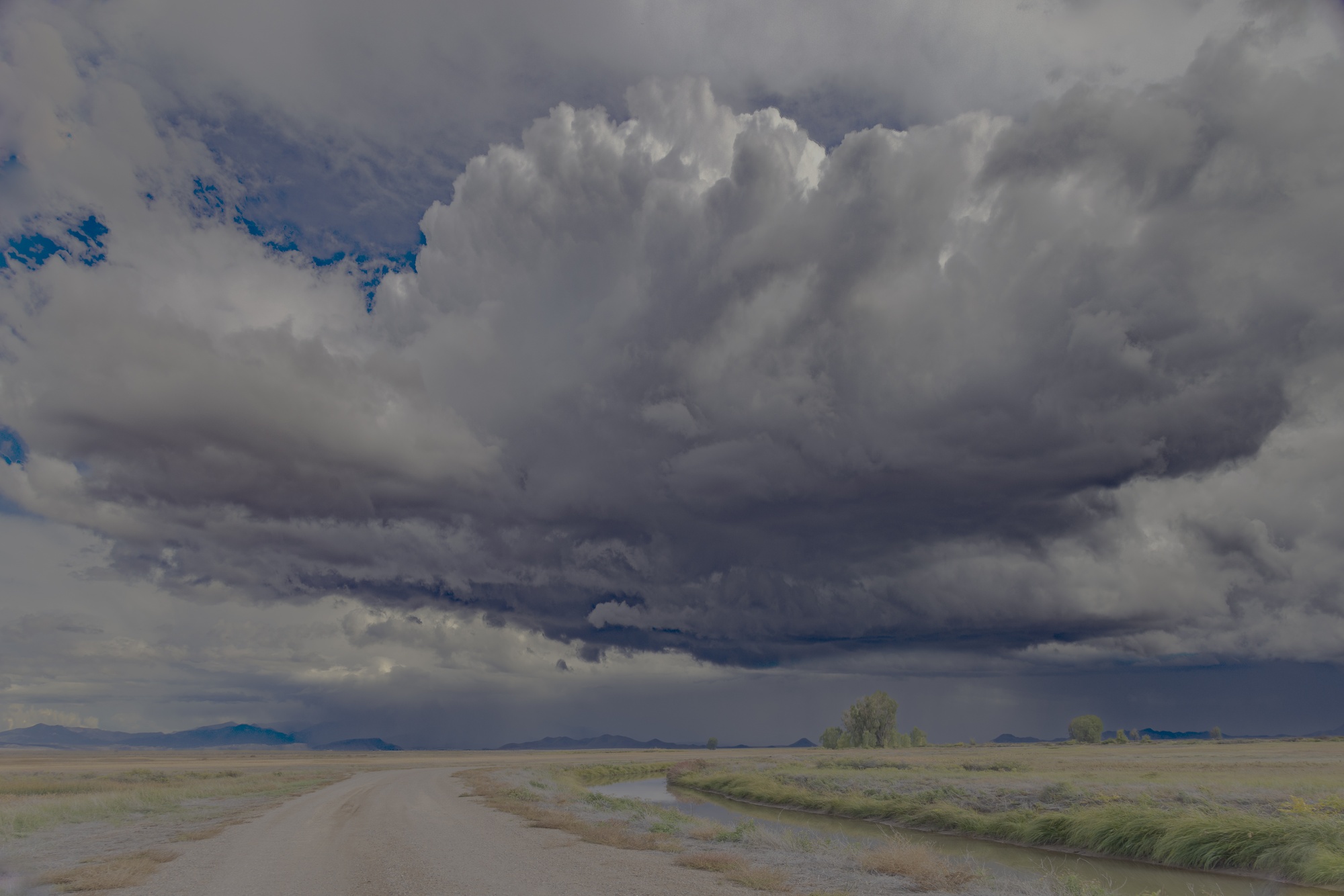 A thunderhead over the Alamosa wildlife refuge, rain falling in the distance beyond a gravel road and an irrigation ditch